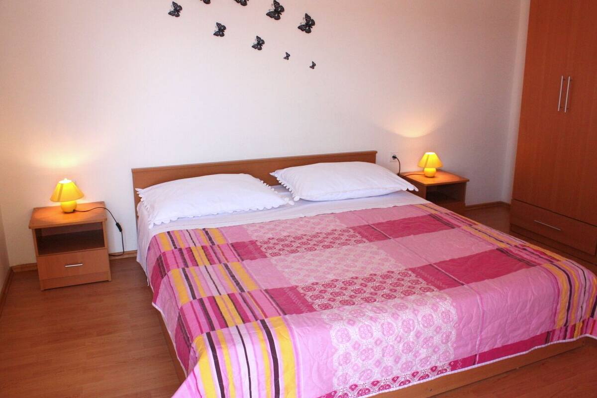 Una cama bien hecha con colcha rosa y blanca, dos lámparas de noche, una mesita de noche de madera y una pared decorada con mariposas.
