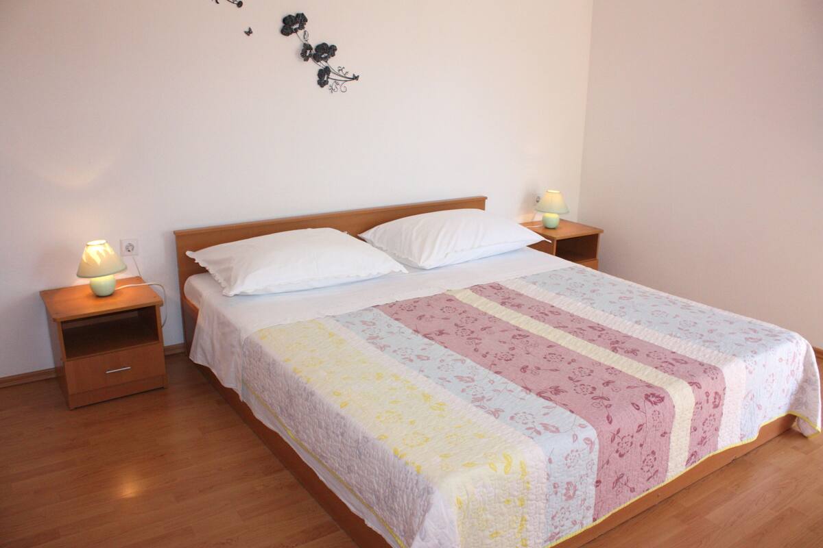 Una cama bien tendida con una colcha de estampado floral, dos mesitas de noche con lámparas y un adorno en la pared.