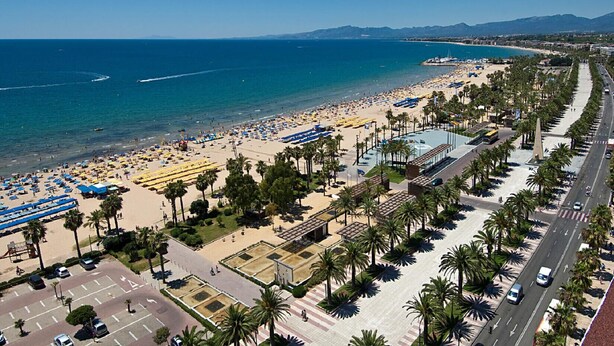 Un paysage urbain côtier avec une plage, une promenade bordée de palmiers et une vue dégagée sur l’océan.