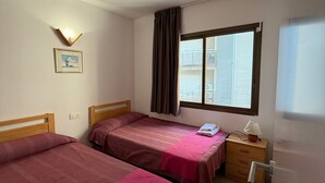 A room with two beds, a nightstand with a lamp, a window with blinds, and a wall-mounted light fixture.