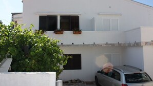 A white two-story building with a car parked in front. There are two windows with shutters, and a small balcony with potted plants.