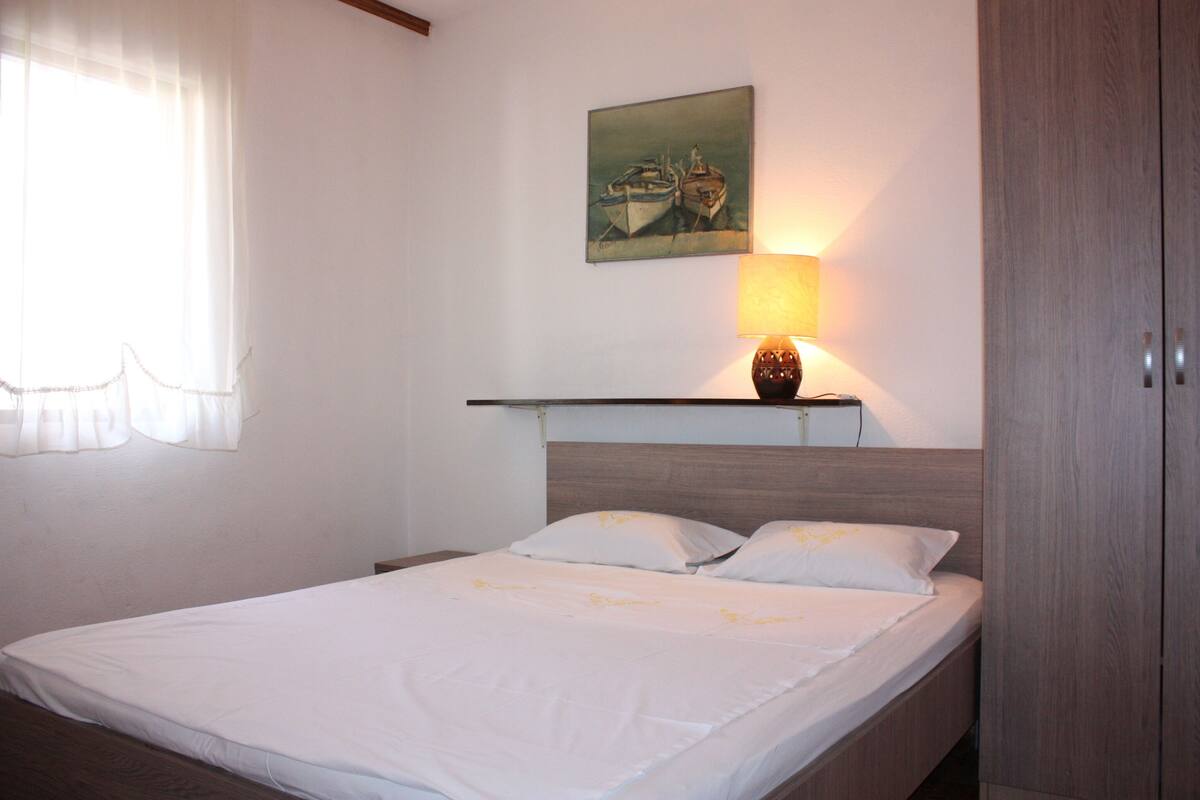 A neatly made bed with white linens, a painting of boats on the wall, and a bedside lamp.