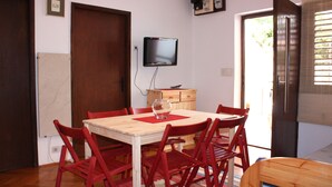 A dining area with a wooden table and red chairs, a TV mounted on the wall, and a door leading outside.