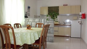 Ein Essbereich mit Tisch für vier Personen, umgeben von Holzsesseln. Im Hintergrund ist eine Küche mit Kühlschrank, Herd und Schränken erkennbar.