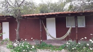 A rustic building with a red exterior, a hammock hanging over the entrance, and a garden with white flowers. - Descanso puntano (Carpintería)