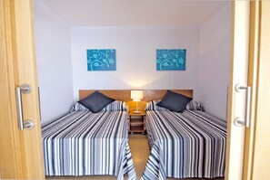 Een kamer met twee bedden, elk voorzien van gestreepte lakens, een houten hoofdbord en een nachtkastje met een lamp. Boven de bedden hangen twee blauwe muurdecoraties met bloemen.
