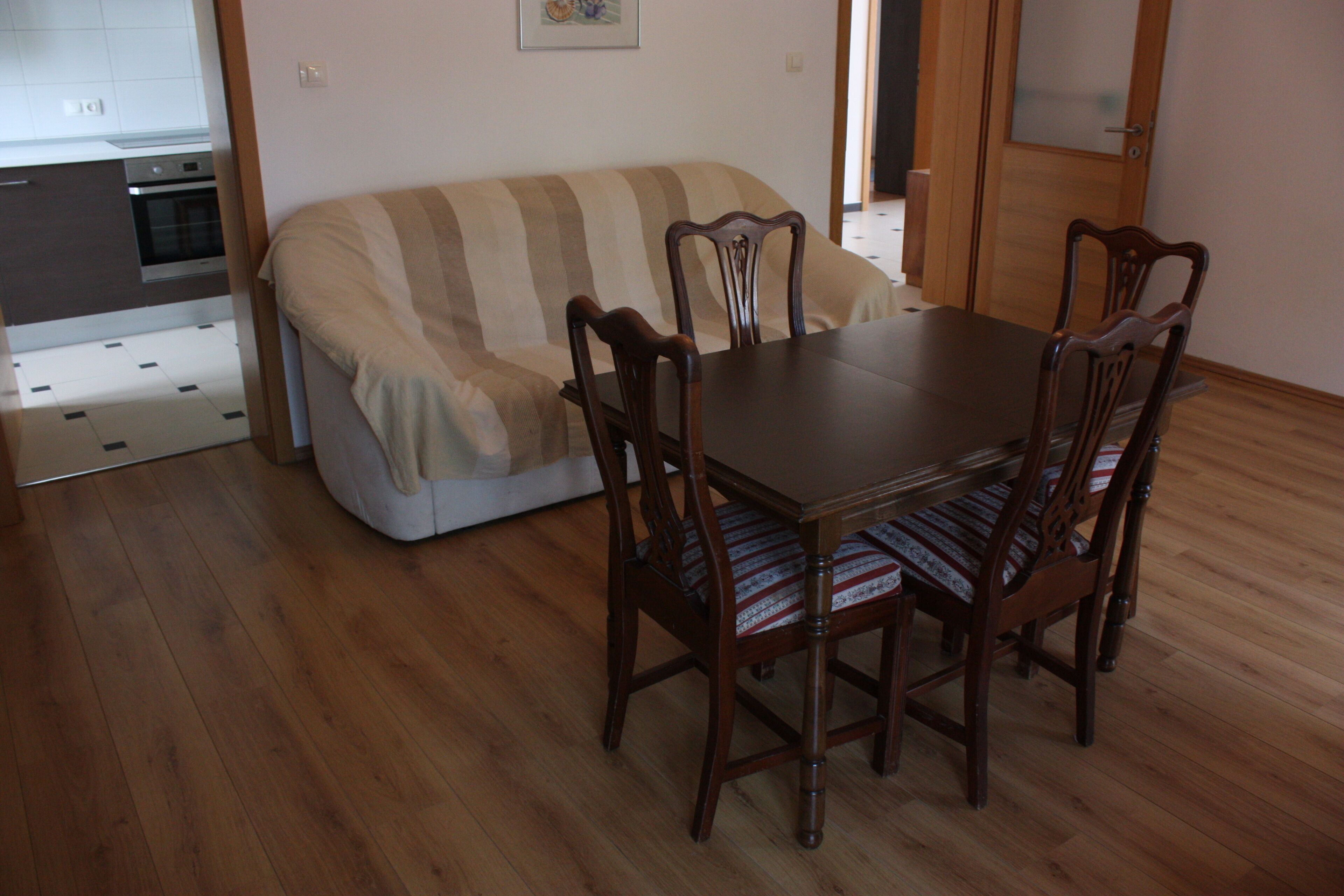 A dining area with a wooden table and four chairs, a striped sofa, and a kitchen in the background.