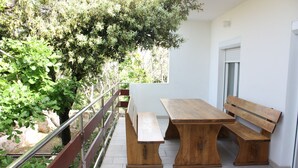 A wooden bench and table set on a balcony with a railing, overlooking a garden with trees and a stone wall. - Pintar Apartment (Pag)