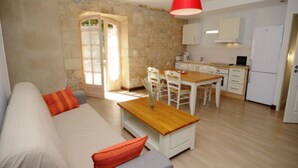 Una sala acogedora con un sofá, una mesa de centro de madera y un comedor con mesa y sillas. Se ve una cocina con gabinetes y electrodomésticos blancos al fondo.