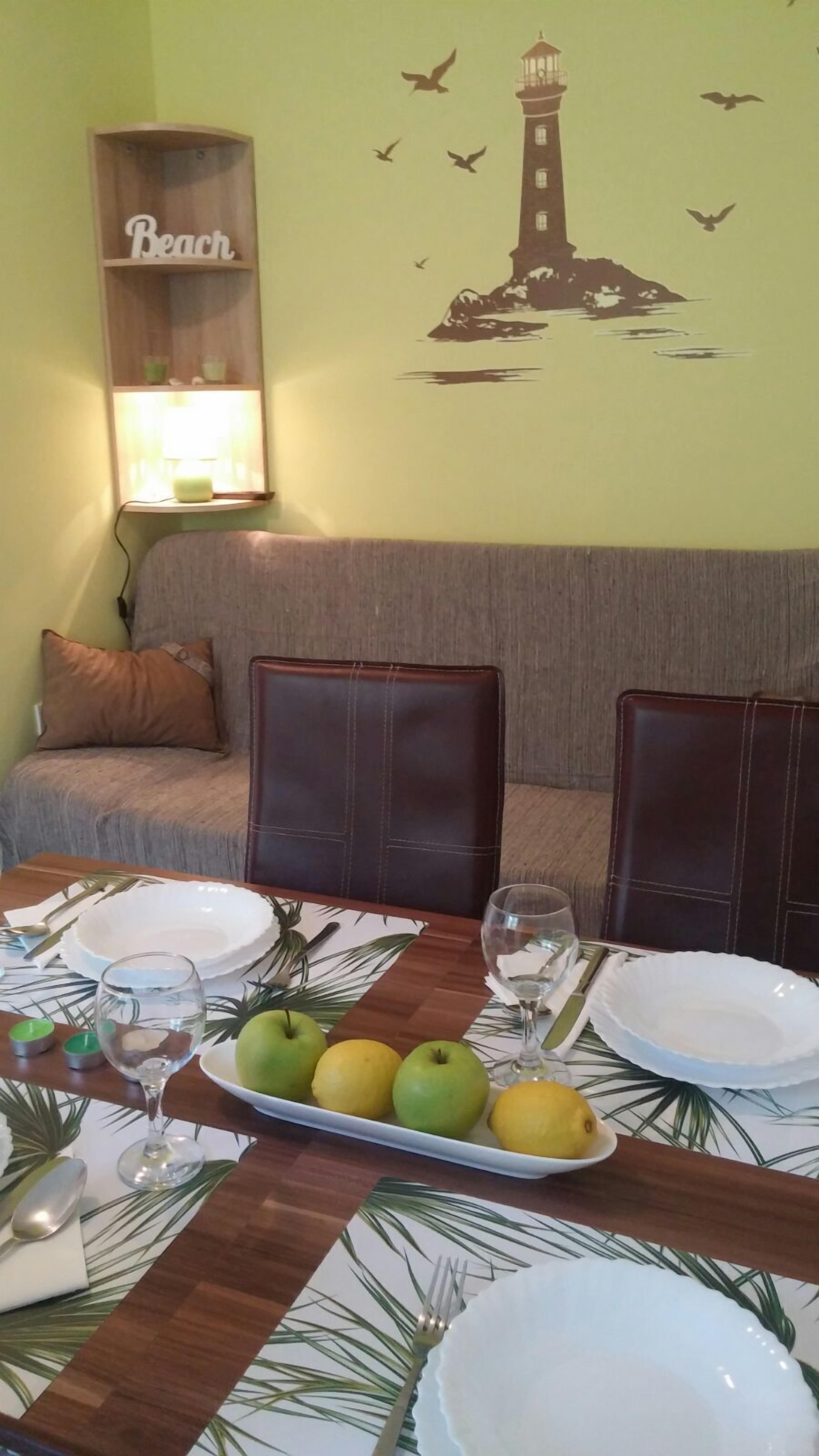 Un comedor con una mesa preparada para dos personas, que incluye un plato de fruta, una copa de vino y una vela. Se aprecia un adorno en la pared con la imagen de un faro.