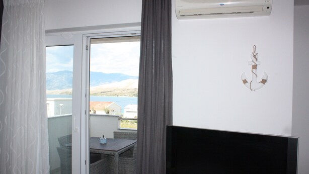 Una stanza con televisione, un tavolo da pranzo con sedie, vista su montagne e un corpo d'acqua attraverso una porta vetrata, e una parete bianca con un ornamento appeso.