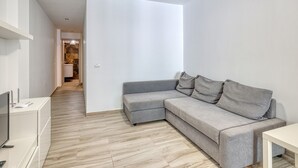 Una sala moderna con un sofá gris, una pequeña mesa blanca y un televisor sobre un mueble. El cuarto tiene piso de madera y paredes blancas.