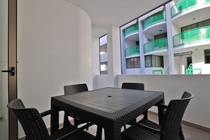 Un balcón con una mesa y sillas, vista a un edificio con balcones verdes y una puerta a la izquierda.