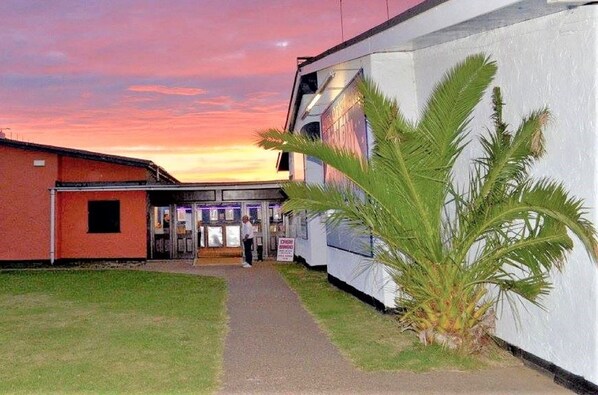 A building with a pathway leading to a door, a palm tree in front, and a sunset in the background.
