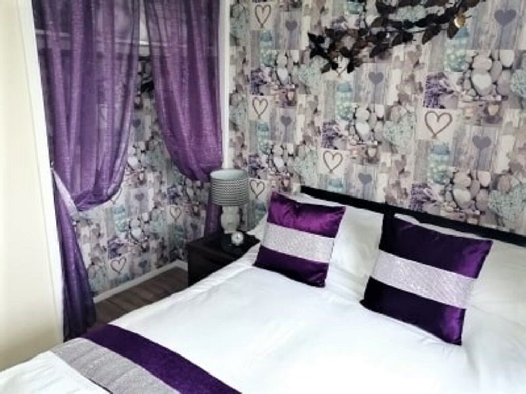Une chambre à coucher avec un lit, des rideaux violets, une table de chevet et un mur orné d’un motif floral.