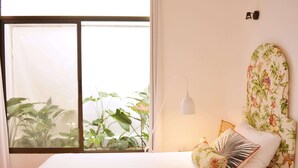 Een slaapkamer met een bed, een raam met planten, een wandlamp en een bloemistisch hoofdeinde.