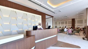 A modern reception area with a wooden reception desk, a digital display, and a seating area with tables and chairs. - Be Inn Hotel (Seeb)