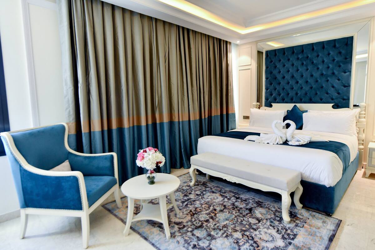 A hotel room with a large bed, a blue armchair, a small round table with a vase of flowers, and a patterned rug.