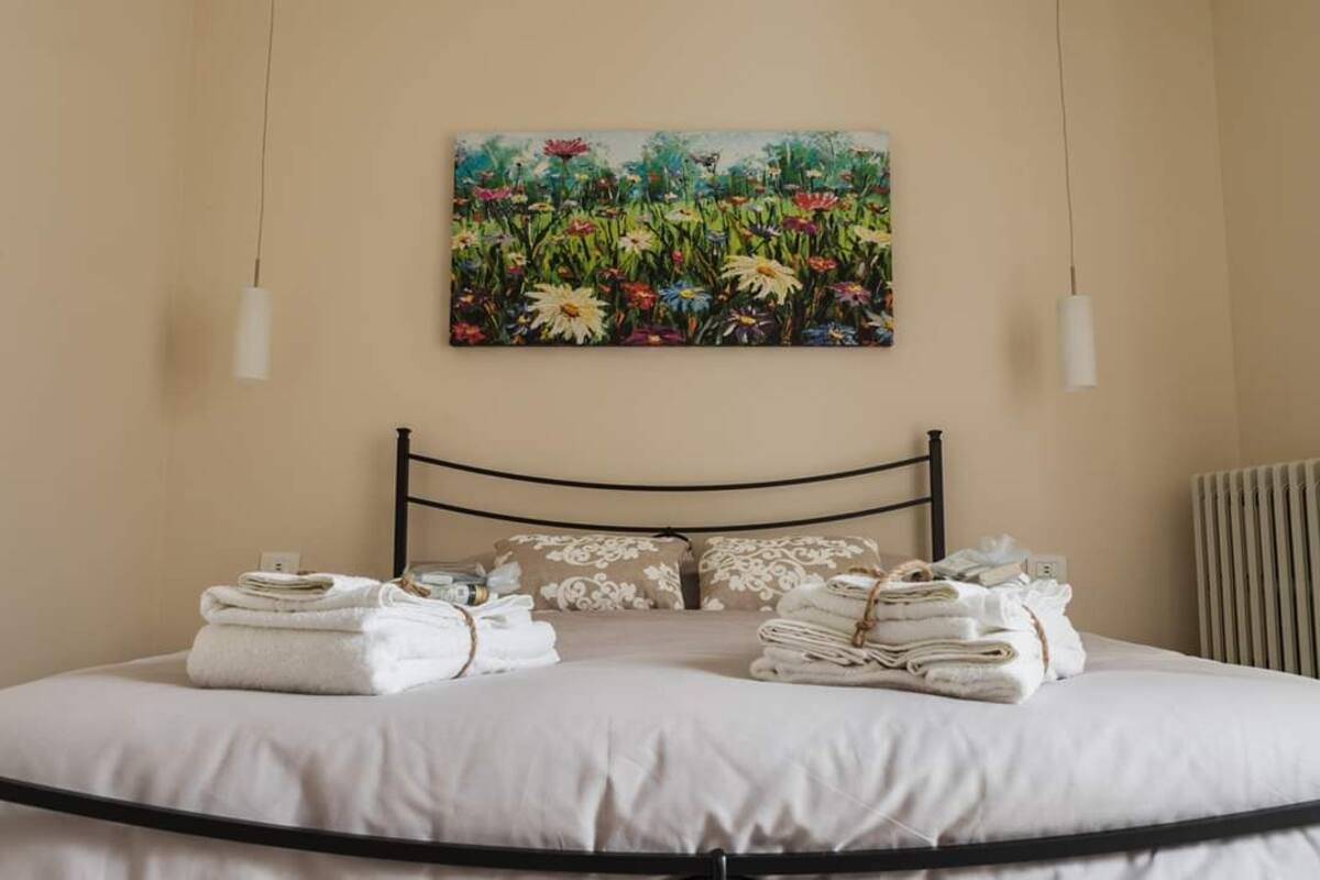 A neatly made bed with folded towels, a floral painting above it, and a radiator on the wall.