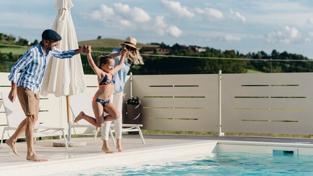 A swimming pool area with lounge chairs and umbrellas, surrounded by a fence and greenery in the background.