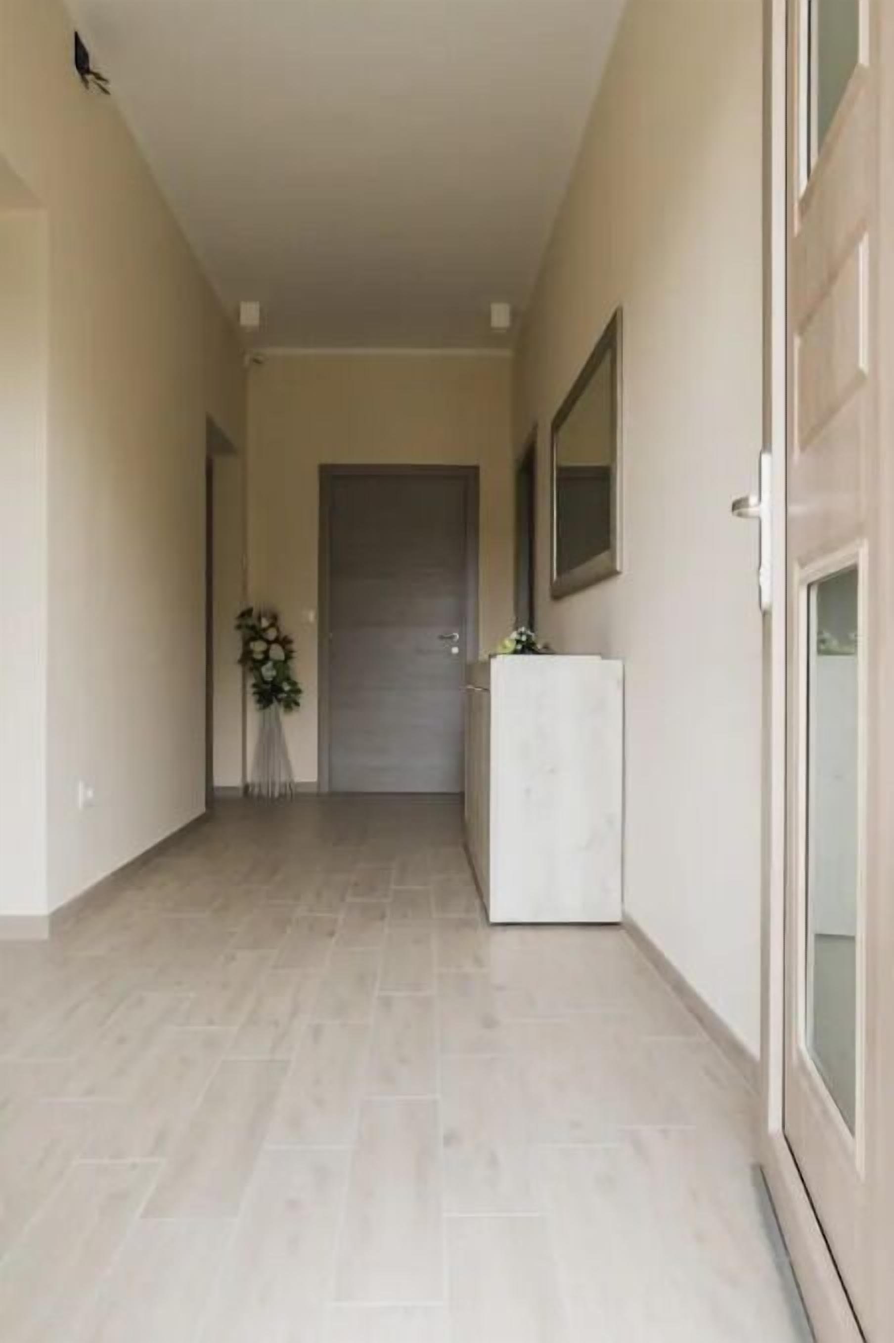 Un corridoio con una porta in fondo, un tavolino bianco con un vaso di fiori e uno specchio sulla parete.