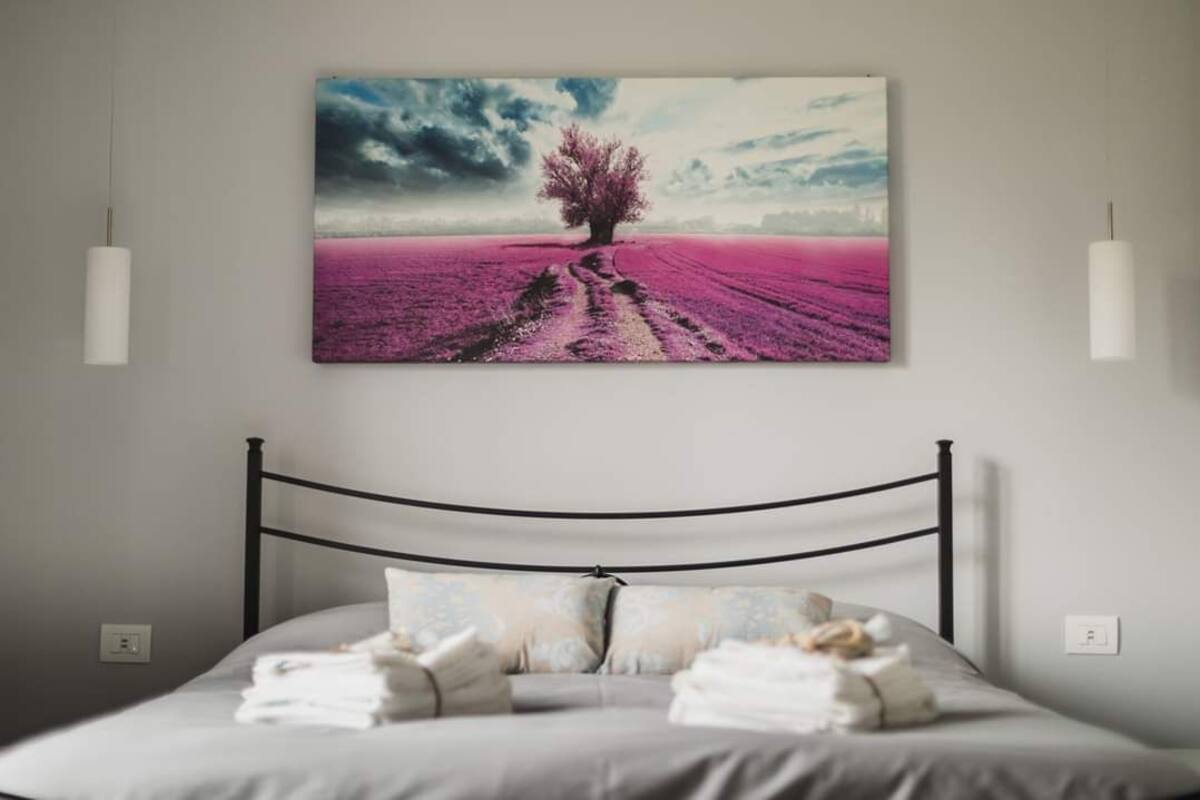 A bedroom with a bed, pillows, and a wall-mounted painting of a tree in a field.