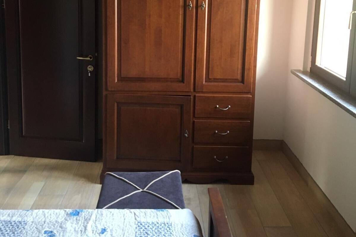 A bedroom with a wooden wardrobe, a bed with a patterned bedspread, and a window with daylight.