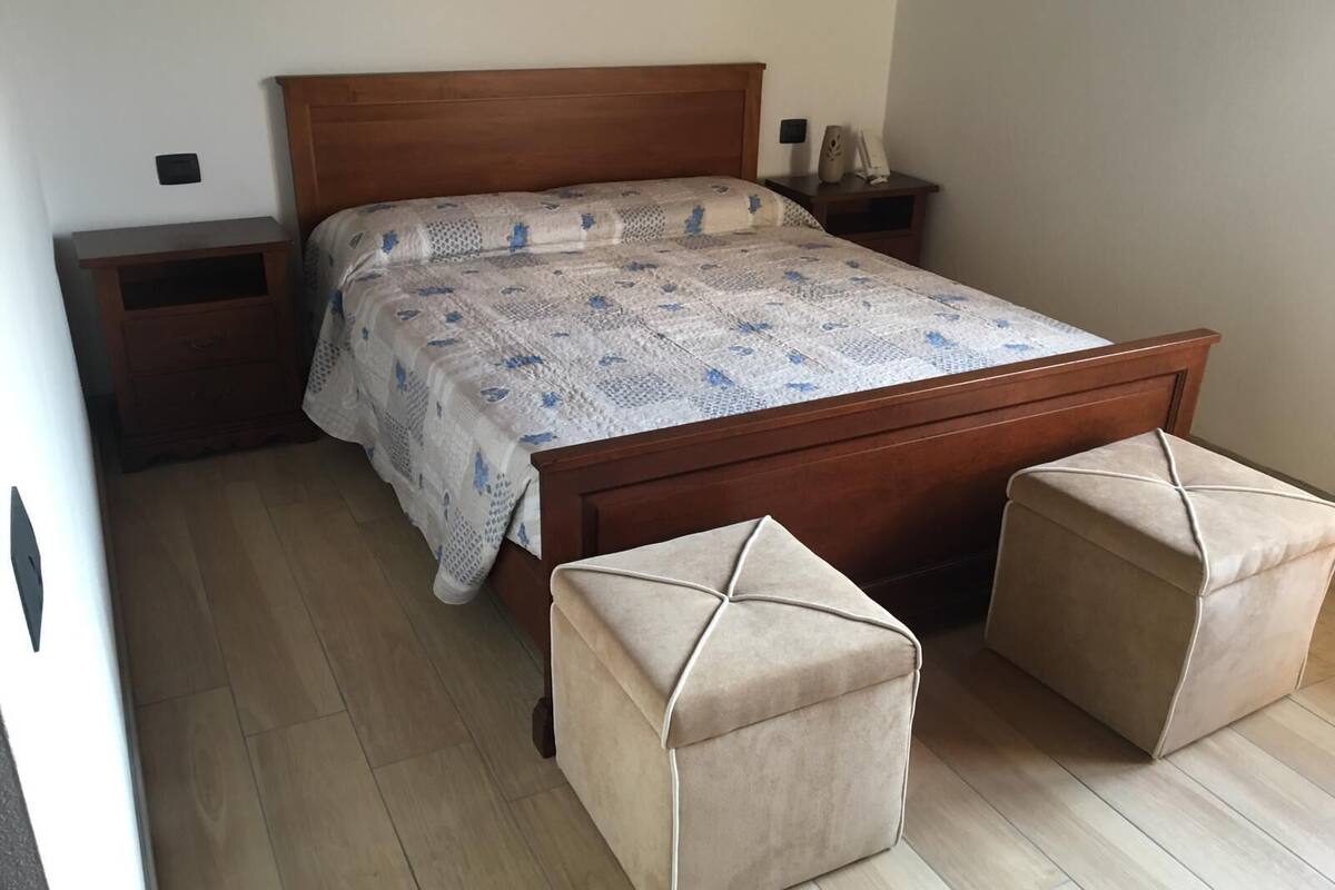 A bedroom with a wooden bed, a nightstand, a bedside table, and two ottomans.