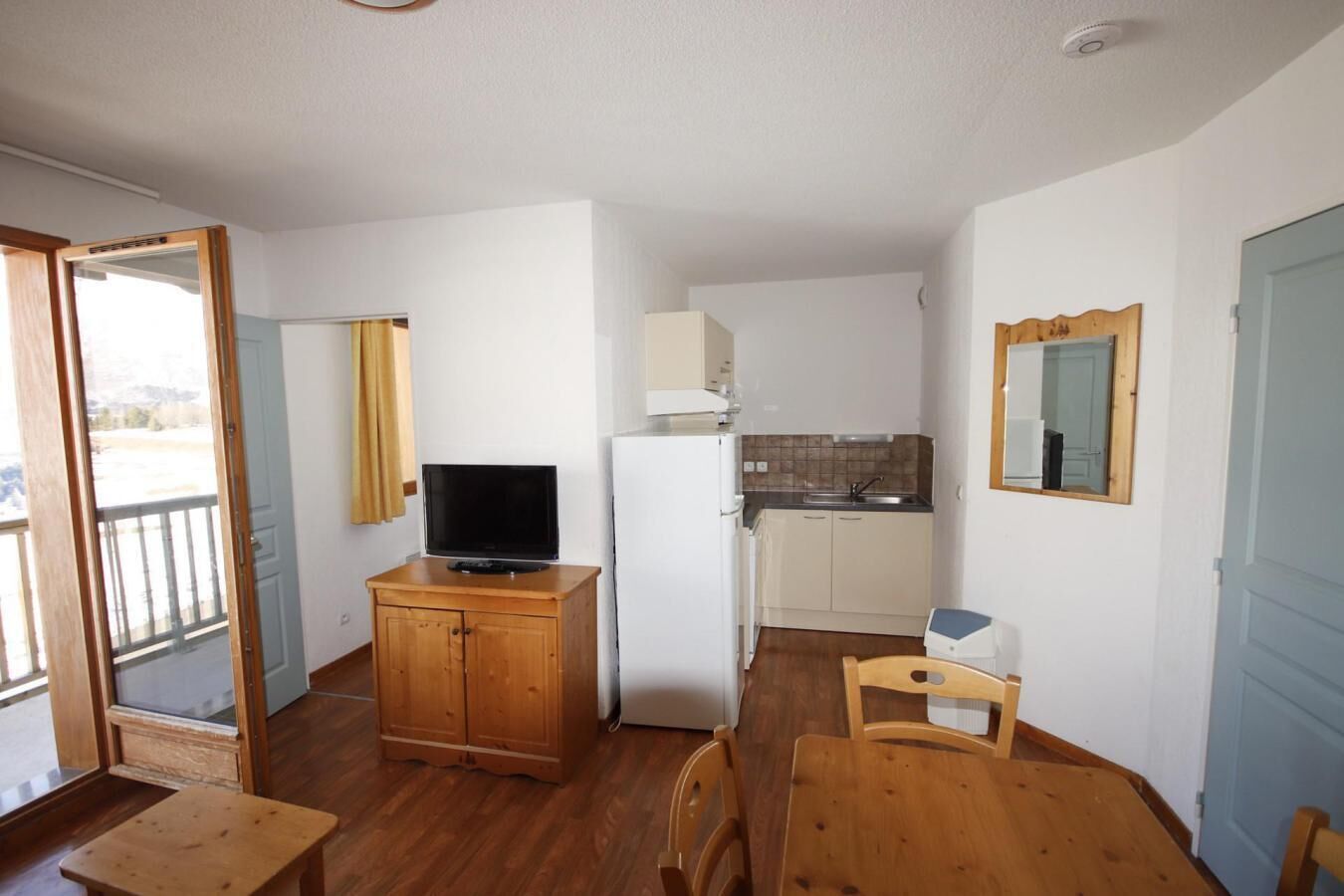 A compact room with a wooden cabinet, a television, a small kitchen area, and a dining table with chairs.
