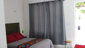 Un dormitorio con cama, ventilador de techo, ventana con cortinas, un taburete pequeño y una silla roja.
