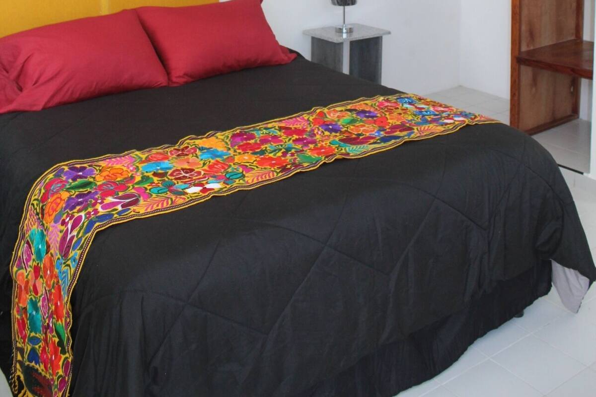 A bedroom with a bed, a red pillow, a colorful blanket, a wooden wardrobe, and a wall with a large painting.