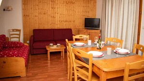 Een knus interieur met houten vloer, rode bank, eettafel voor twee personen en een televisie op de achtergrond.