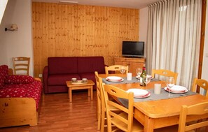 Ein gemütliches Zimmer mit Holzboden, einem roten Sofa, einem Esstisch für zwei Personen und einem Fernseher im Hintergrund.