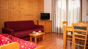 Een woonkamer met een rode bank, een televisie op een houten kast, een eettafel met stoelen en een raam met gordijnen.