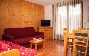 Ein Wohnzimmer mit einem roten Sofa, einem Fernseher auf einem Holzschrank, einem Esstisch mit Stühlen und einem Fenster mit Vorhängen.