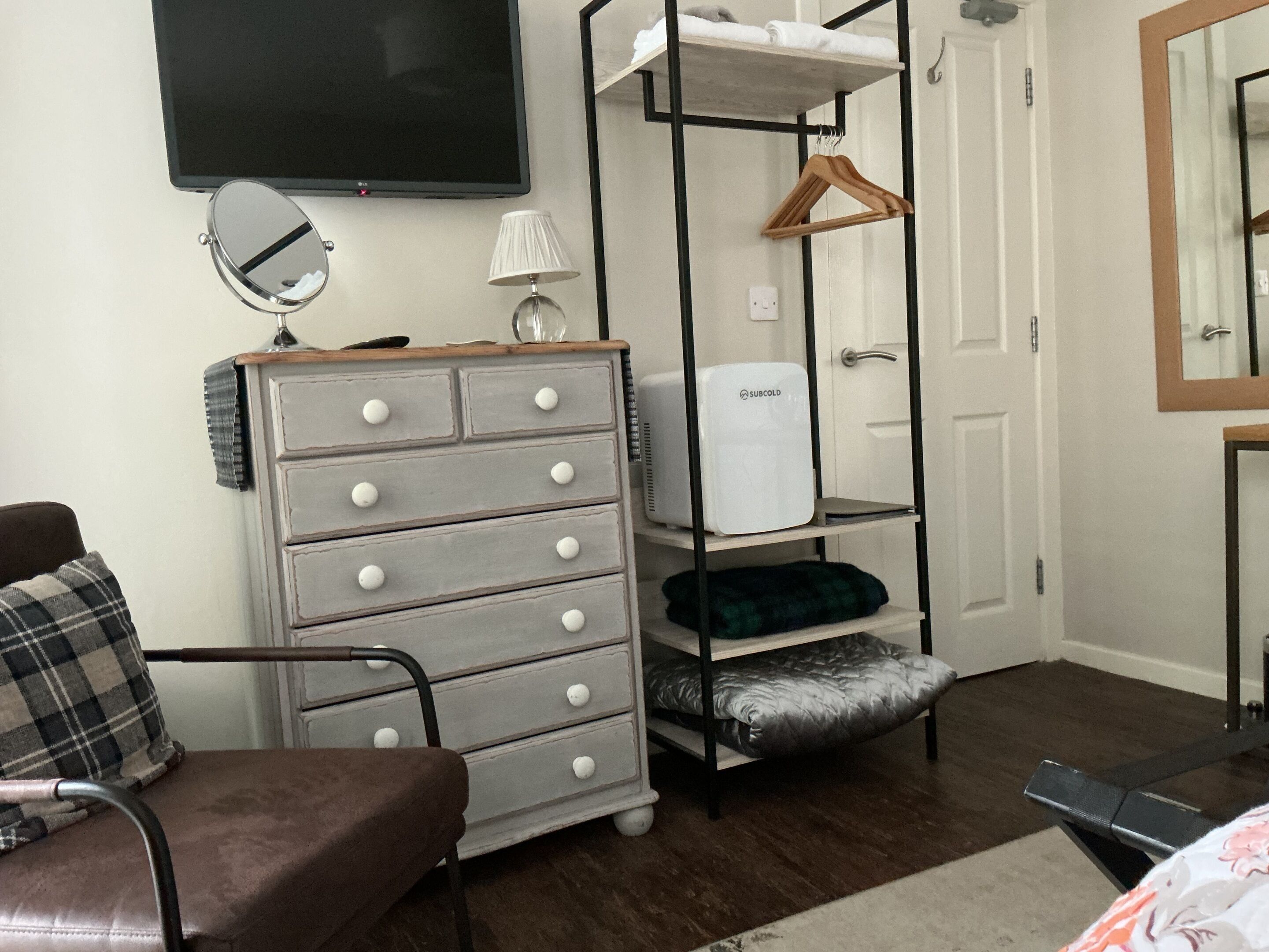A bedroom with a bed, a dresser, a TV, a mirror, a shelf with a heater, and a door.