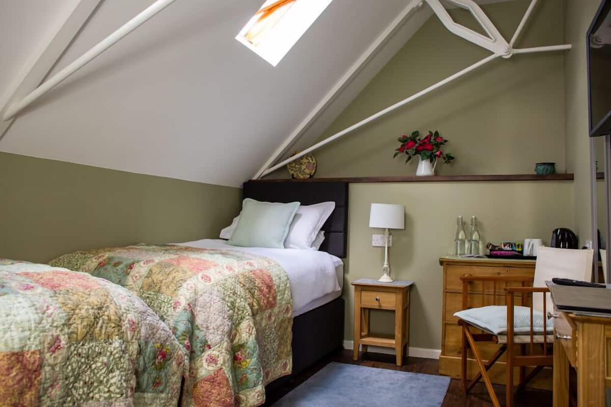 A bedroom with a sloped ceiling, a bed with floral bedding, a bedside table with a lamp, a chair, and a desk with a computer.