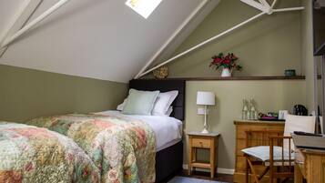 Un dormitorio con techo inclinado, una cama con ropa de cama floral, una mesita de noche con lámpara, una silla y un escritorio con computadora.