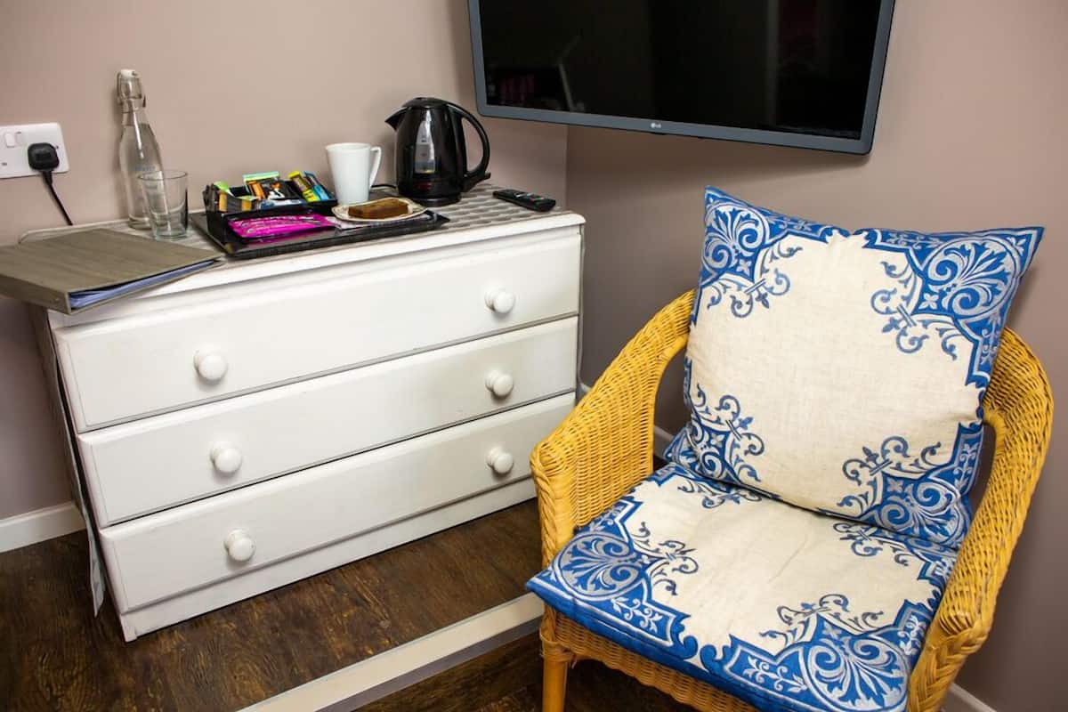 A white dresser with drawers, a wicker chair with a patterned cushion, a flat-screen TV, and a small table with a kettle and cups.