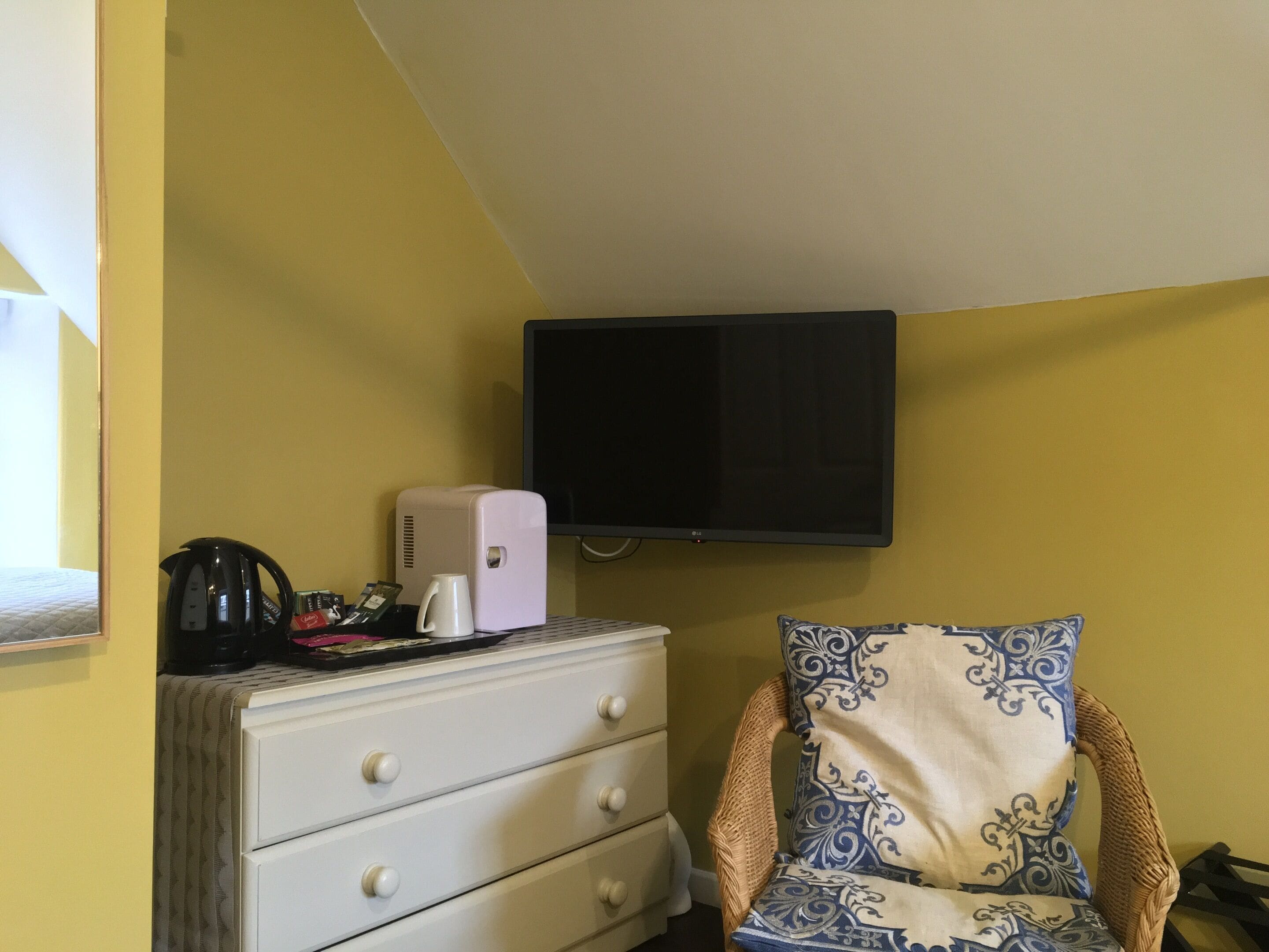 A room with a white dresser, a white electric kettle, a black coffee maker, a white mug, a flat-screen TV, and a patterned armchair.