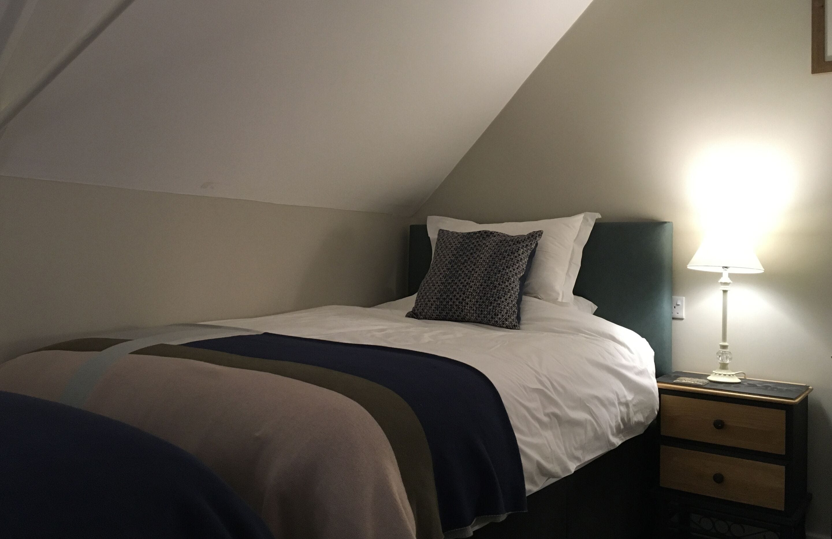 A single bed with a patterned pillow, a bedside table with a lamp, and a small drawer.