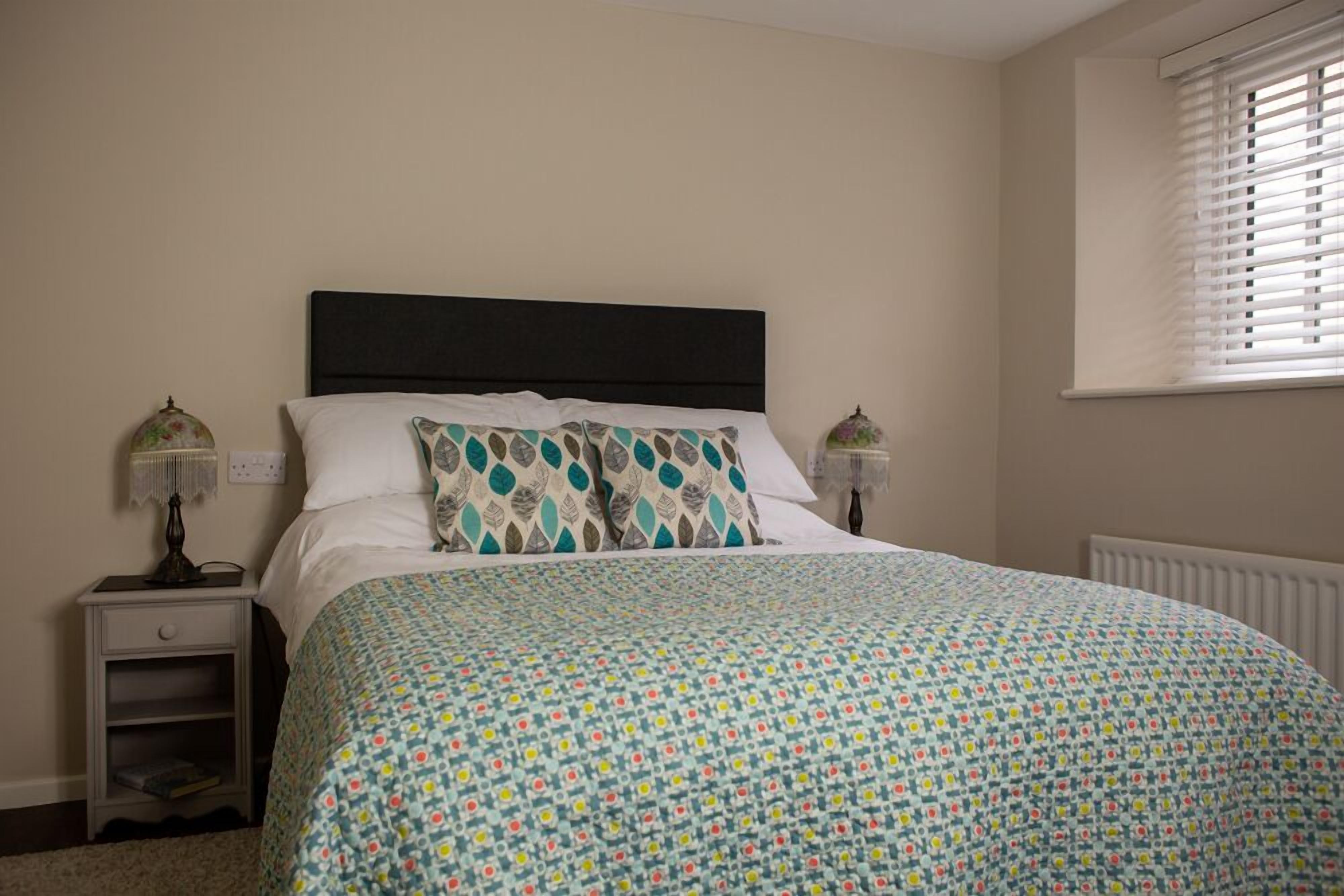 A bedroom with a bed, two bedside lamps, a nightstand, and a window with blinds.