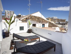 A balcony with a cushioned daybed, a potted plant, and a table with fruit. - MONKÓ BANANA TROPYCAL APARTMENT (Torremolinos)