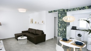 A modern living room with a brown sofa, a round dining table with chairs, a kitchen area with a washing machine, and a ceiling light.