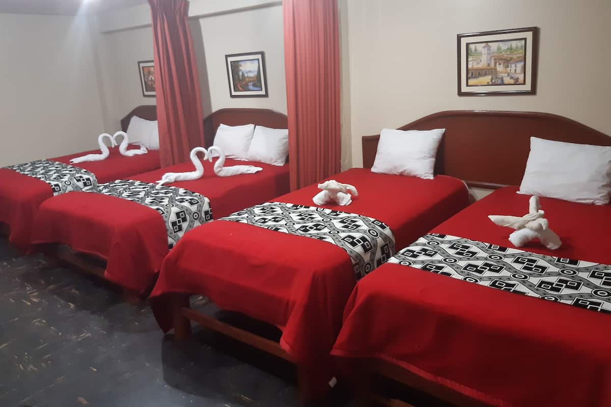 A hotel room with four beds, each with a red bedspread and a patterned pillow. There are two paintings on the walls and a window with red curtains.