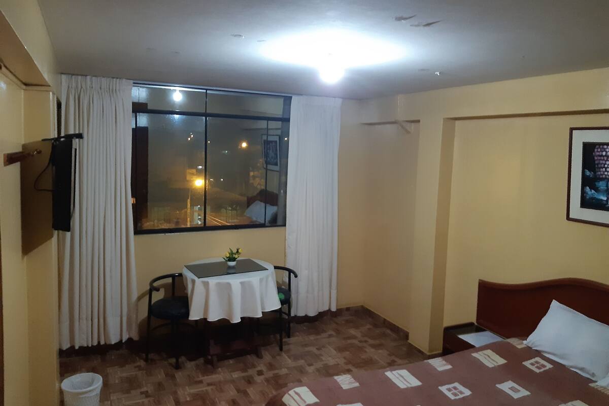A hotel room with a bed, a small table with a vase of flowers, a chair, a window with curtains, and a wall-mounted TV.