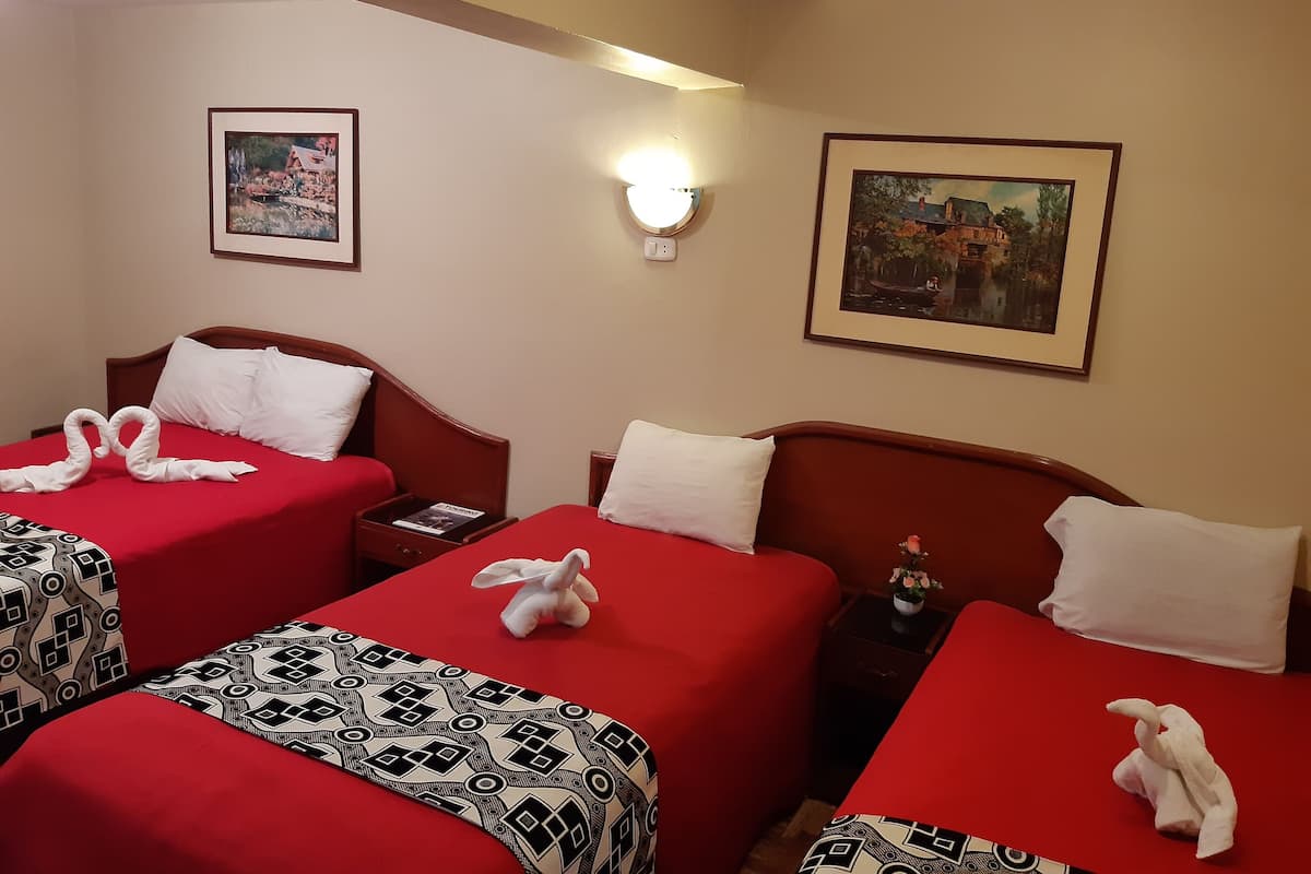 A hotel room with two beds, each with a red bedspread and white pillows. There are framed pictures on the walls, a bedside table with a lamp, and a small potted plant on the nightstand.