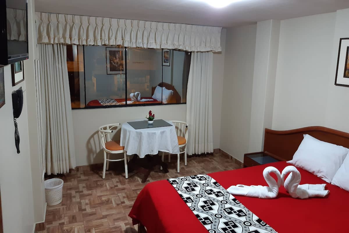 A hotel room with a bed, a small table with a vase of flowers, a chair, a window with curtains, and a wooden headboard.
