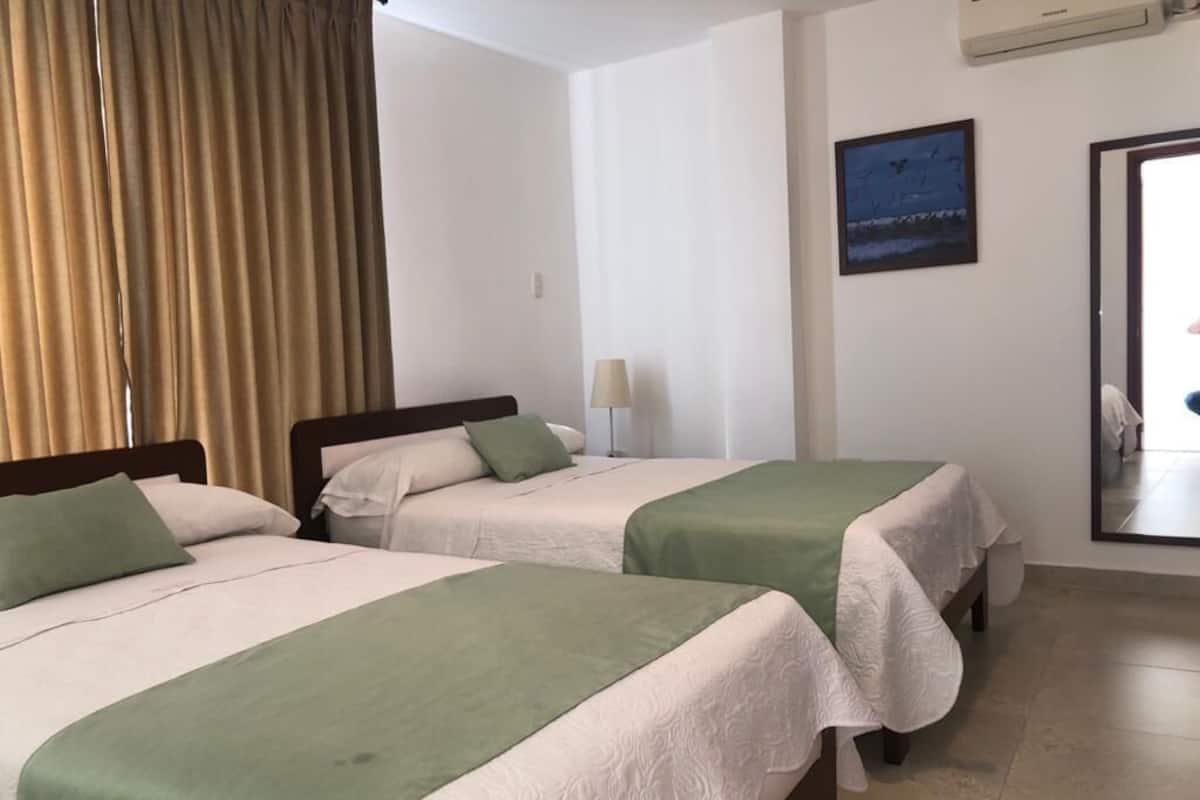 A hotel room with two beds, beige walls, a window with curtains, a lamp, and a framed picture on the wall.