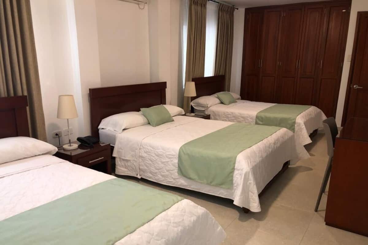 A hotel room with three single beds, each with a green bedspread and white linens. There are wooden headboards, a nightstand with a lamp, and a wardrobe with a mirror.